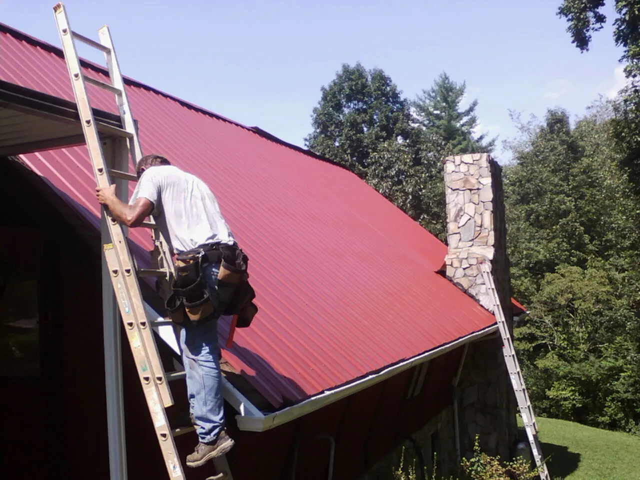 Roofer on a ladder beside a red metal roof and stone chimney — Soffit & Fascia Repair by Roofing Brothers in Tomkins Cove
